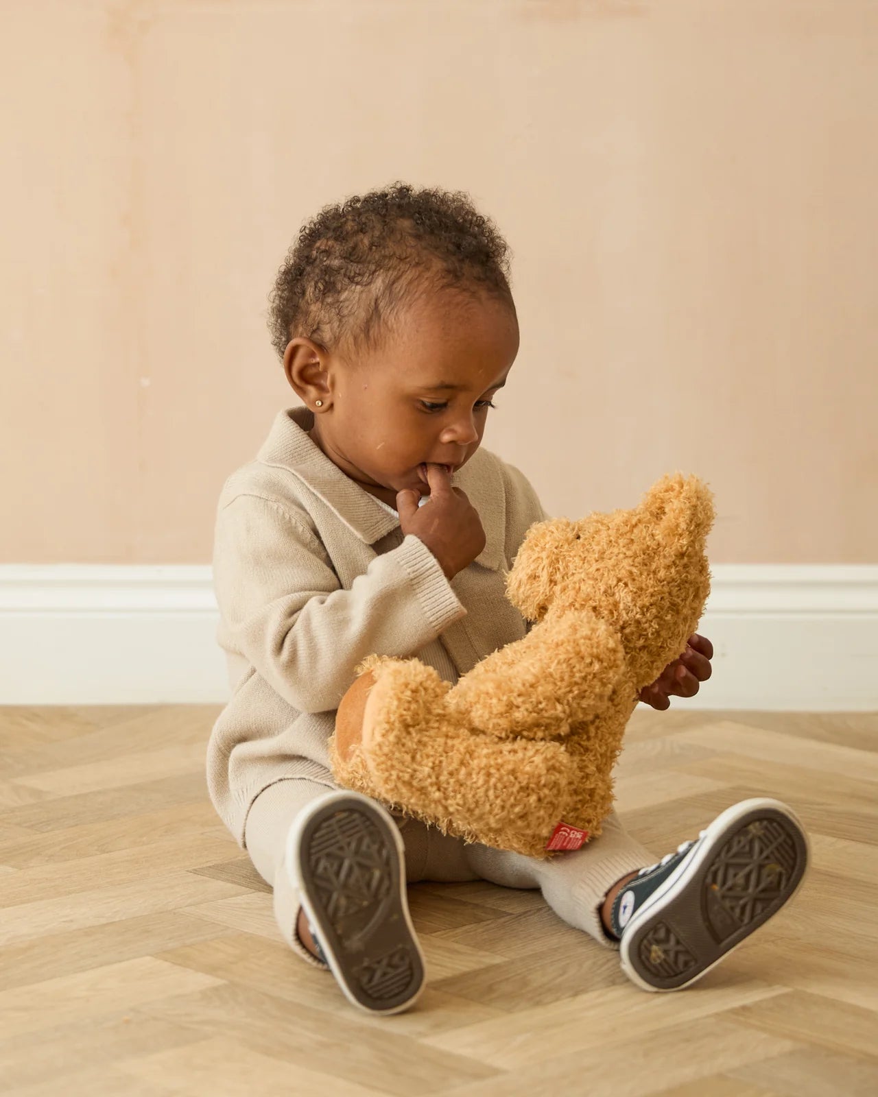 Child sitting on a wooden floor holding a teddy bear against a beige wall.