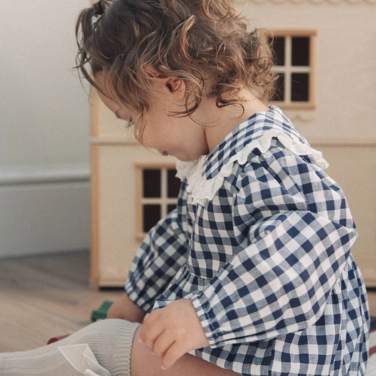 Child in a blue and white checkered outfit sitting on the floor with a wooden dollhouse in the background.