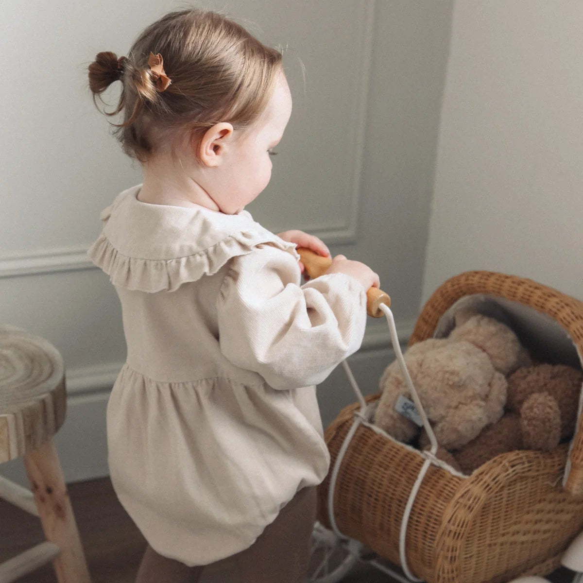 Child pulling a wicker toy pram with teddy bears inside against a neutral wall.