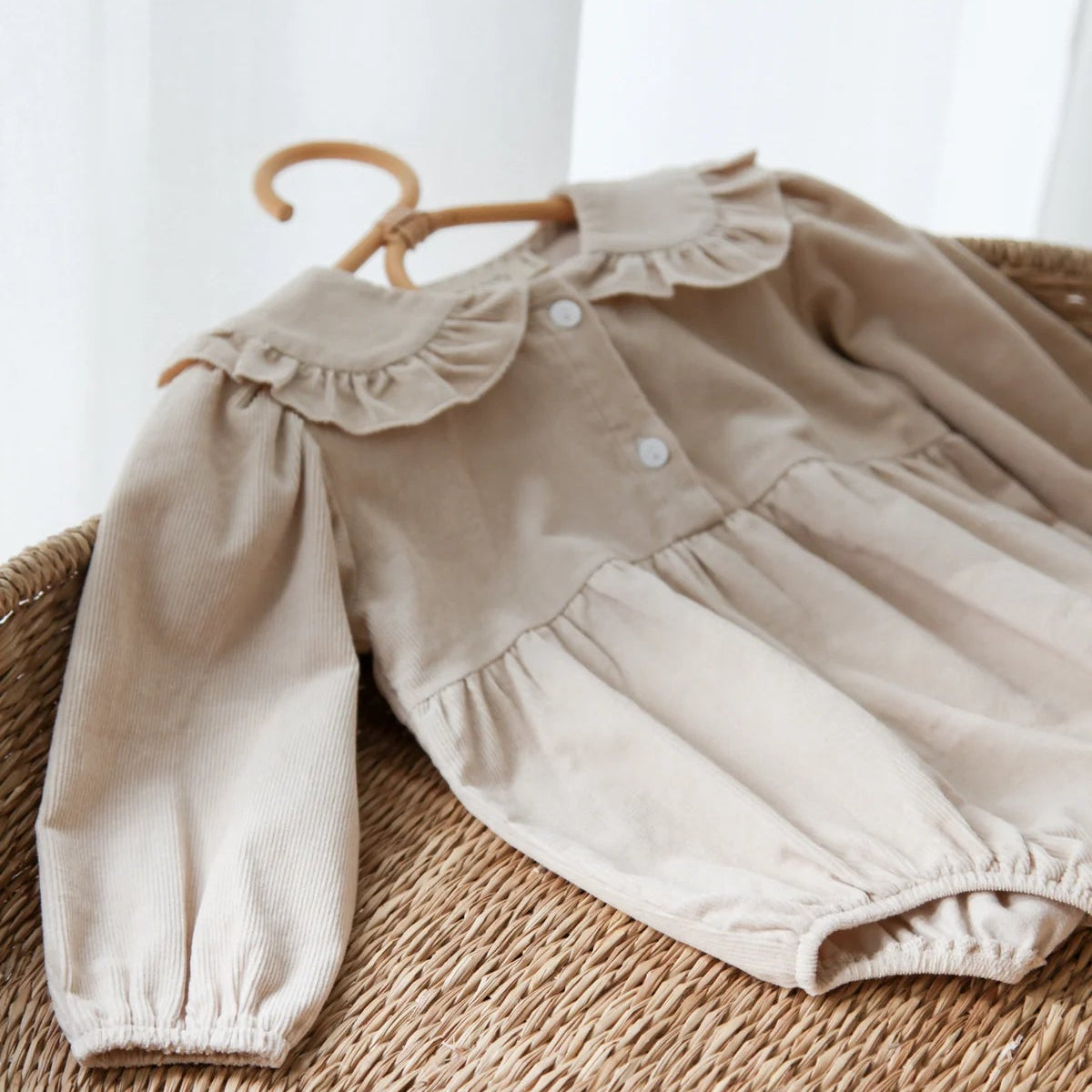 Beige baby outfit with ruffled collar on a wicker basket against a white curtain background