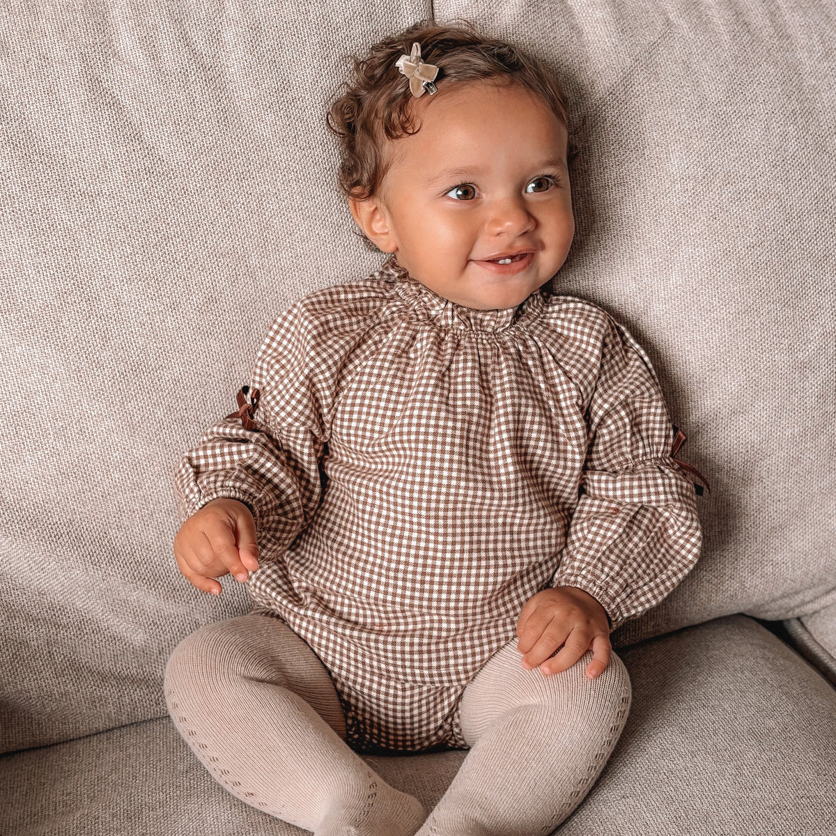 Baby wearing a checkered outfit sitting on a gray couch.