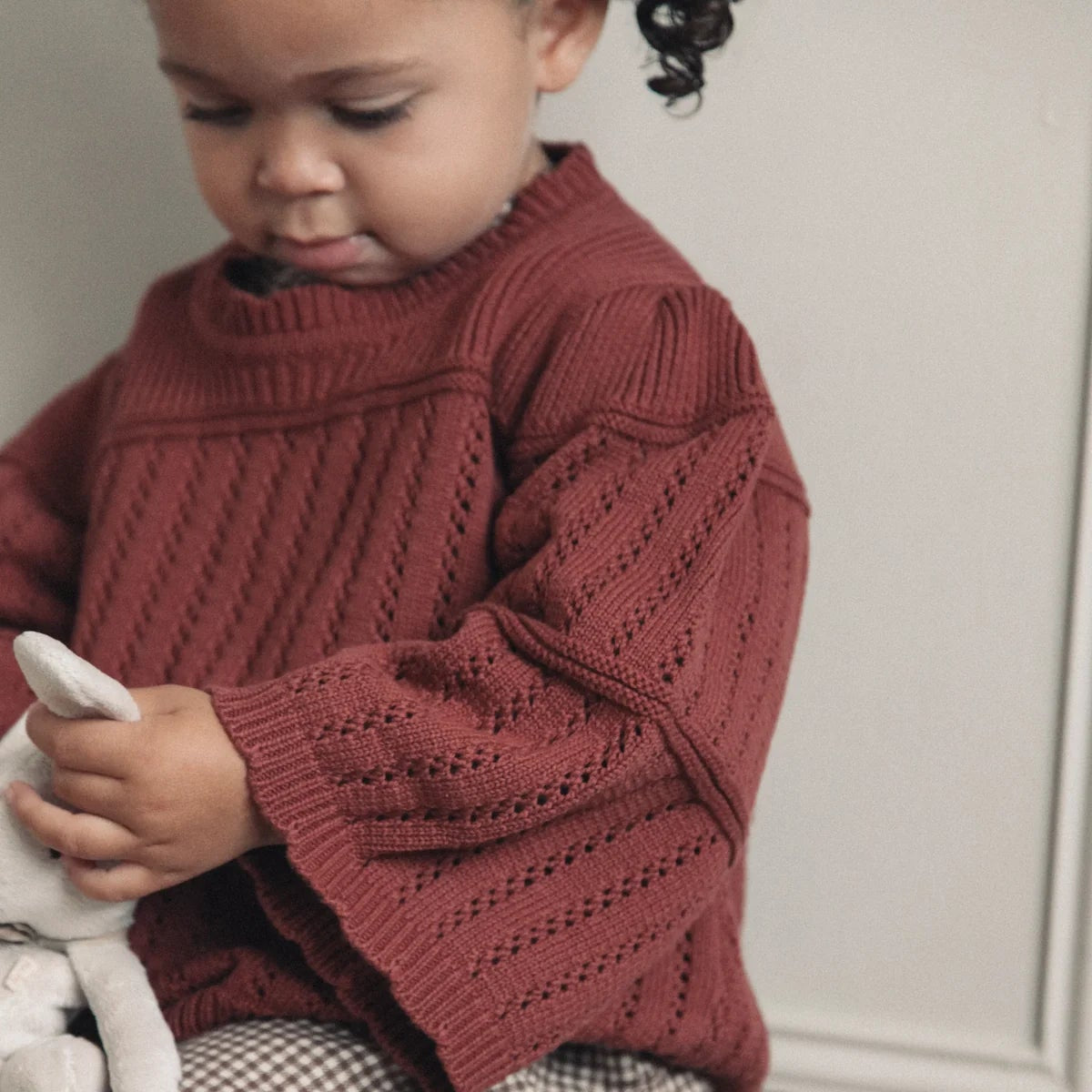 Child wearing a red sweater sitting on a wooden stool holding a stuffed toy.