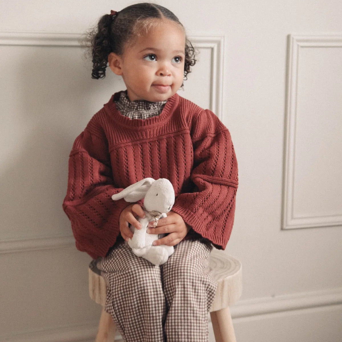 Child wearing a red sweater holding a stuffed animal in a room with white walls.